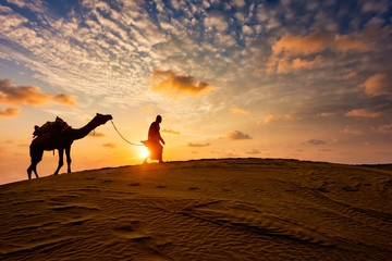 Indian cameleer (camel driver) bedouin with camel silhouettes in sand dunes of Thar desert on...