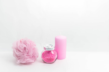 A transparent jar with bath salt with a metal lid, a sponge for bath and a candle stand nearby on a white background. All objects have a light pink color. The concept of items for shower, personal