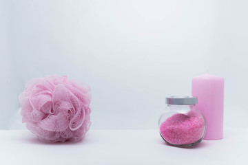 A transparent jar with bath salt with a metal lid, a sponge for bath and a candle stand on a white background. All objects have a light pink color. The concept of items for shower, personal care and