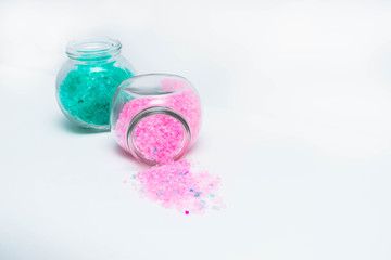 Jars with turquoise and light pink bath salts are on the left side of the frame. Horizontal photo. Objects on a white background. Bank with light pink salts is lie on its side and salt spilled out