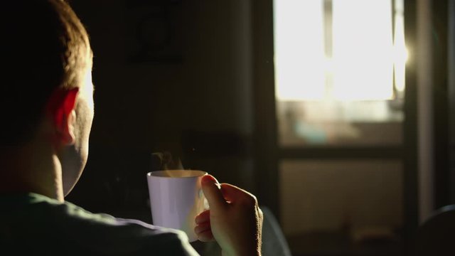 Silhouette of a man with a mug of hot drink, against the backdrop of sunrise, rear view, camera movement