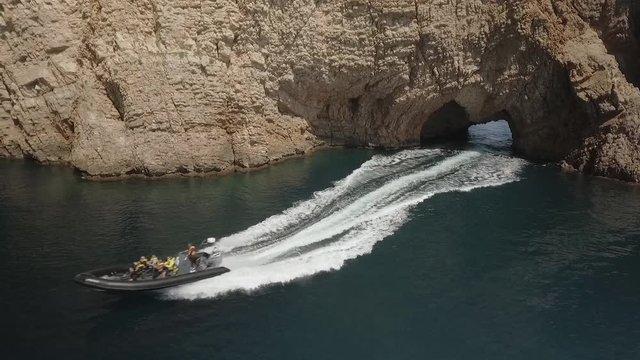 AERIAL DRONE FOLLOWING SPEEDBOAT COMING OUT OF CAVE IN MEDITERRANEAN SEA RED ROCKY CLIFF