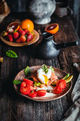 breakfast bowls with poached eggs, vegetables and salmon, oranges and fresh strawberry in plate with mugs of coffee on brown wooden table