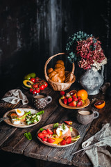 breakfast bowls with poached eggs, vegetables and salmon, oranges and fresh strawberry in plate with mugs of coffee on brown wooden table