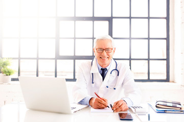 Senior male doctor sitting at desk and working on laptop