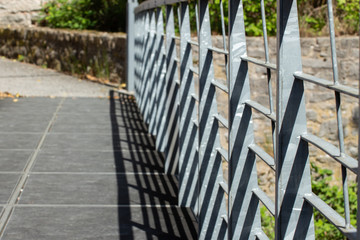 Slanted view of a metal railing on a bridge casting shadows with natural stone walls in the background, focus on foreground