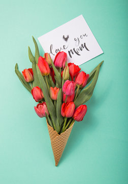red  beautiful tulips in an ice cream waffle cone with card Love you mom  on a color blue background. Conceptual idea of a flower gift. Spring mood