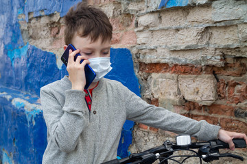 A serious young European boy in a medical mask is talking on the phone with a friend and relative on the street near the blue wall.