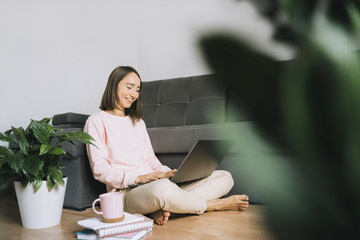 Happy young woman sitting on the floor near couch and using laptop. Frelance work from home in quarantine concept.