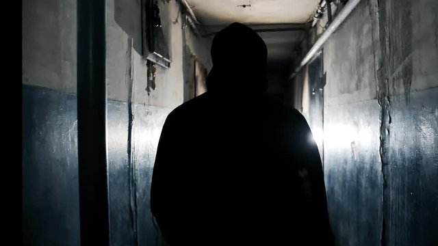 The Camera Follows Unrecognizable Man Going Along the Walkway in the Dark Dirty Shabby Apartment Building. Rear View of Silhouette of a Criminal Man in Black Jacket