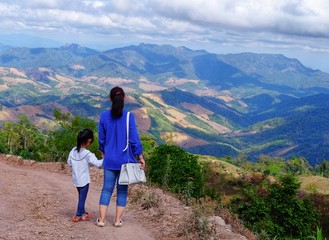 A back view of mother and daughter walking on a path down the mountain with beautiful mountain range and valley in the distance.