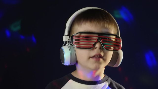 Close-up in a dark room with disco lights, a little boy listens to music in wireless headphones and stylish neon glasses, sings a song and shakes his head