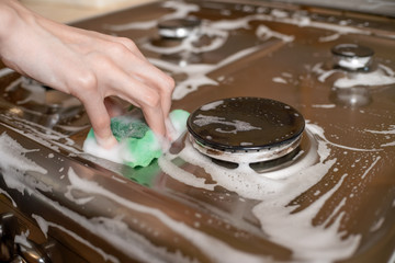 woman's hand washes a gray stainless steel gas stove with a green foam sponge, close up. House cleaning