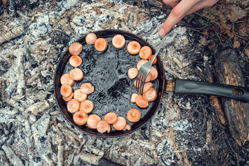 Sausages are fried in a pan on coals in the forest. The girl's hand turns the sausage with a fork in a pan on the coals. Breakfast in nature. Picnic.