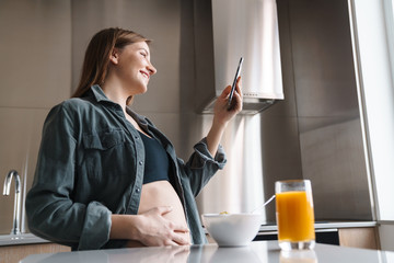 Photo of joyful pregnant woman using cellphone while having breakfast