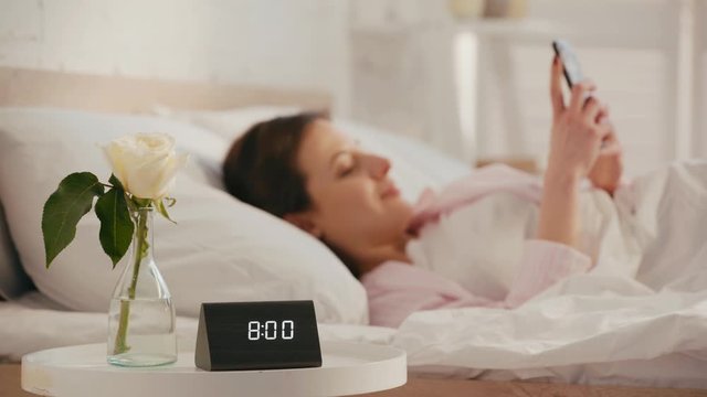 Clock and rose on table near smiling girl using smartphone on bed at morning