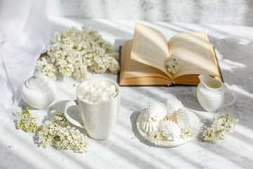 Fototapeta na wymiar Still life vase with a bouquet of white lilac, a Cup of coffee with marshmallows, a plate of meringue, a sugar bowl and a milk jug , an old book. Postcard good morning.