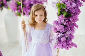 Beautiful little girl of preschool age in a delicate dress sitting on a swing decorated with flowers of lilac.