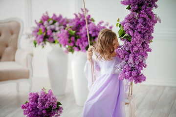 Beautiful little girl of preschool age in a delicate dress sitting on a swing decorated with flowers of lilac.