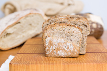 Wholegrain bread slices on chopping board. Closeup shot. Heap of loafs and buns in background. Bakery or healthy breakfast concept