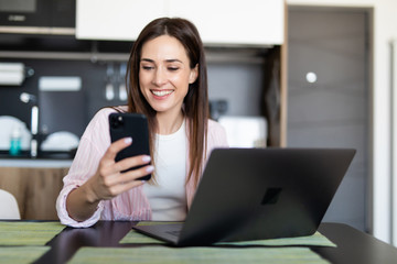 Young woman scroll phone while using laptop at table indoors