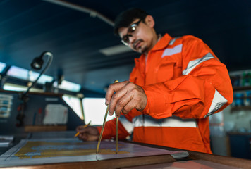 Filipino deck Officer on bridge of vessel or ship wearing coverall during navigaton watch at sea . He is plotting position on chart