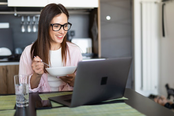 Young cheerful woman using on laptop computer and eating breakfast in the kitchen