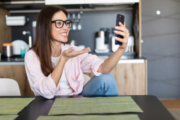 Young smiling cheerful woman at home kitchen using social media apps on phone for video chatting and stying connected with her loved ones. Stay at home, social distancing lifestyle.