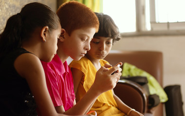 Three multi ethnic kids or siblings busy in playing games on mobile at home - concept of childrens mobile video game addiction, using technology, internet on smartphone.