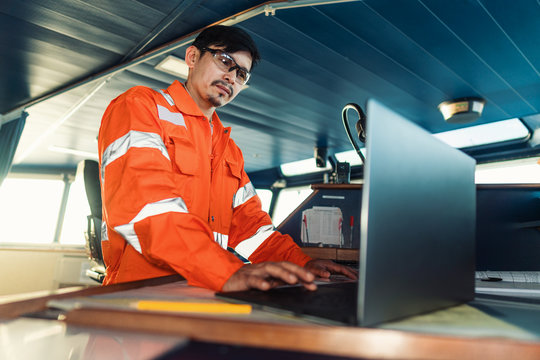 Filipino deck Officer on bridge of vessel or ship wearing coverall during navigaton watch at sea . He is using laptop, electronic paperwork at sea, concept of reporting