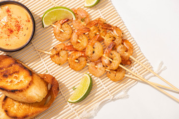 top view of prawns on skewers with grilled bread, lime and sauce on bamboo mat on white background