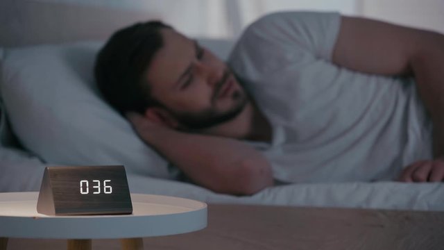 Selective focus of man suffering from insomnia near clock on bedside table