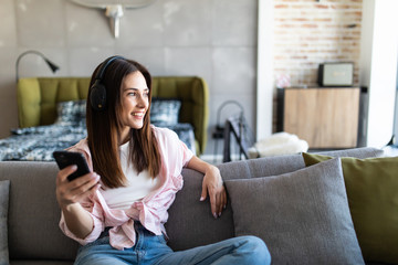 Young woman resting on sofa wearing casual home clothes wireless headphones enjoy weekend free time using online application on cellphone listens music