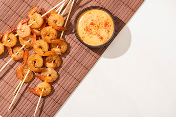 top view of tasty prawns on skewers with sauce on bamboo mat on white background