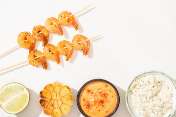 top view of delicious fried prawns on skewers with garlic, lime and sauces on white background