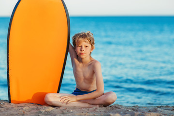 A blonde boy with a surfboard sitting on sand, on a  blue sea background.