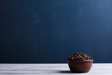 Obraz na płótnie Canvas Coffee beans in clay bowl on white wooden table against dark blue wall background. Closeup, copy space, side view. Arabica seeds, roast, caffeine concept