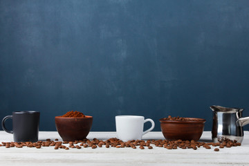 Espresso cups, metal Turkish pot, roasted Arabica beans in clay bowls, coffee powder on while table against dark blue wall. Side view, copy space. Coffee shop, morning, baristas workplace  concept