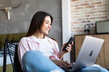 Portrait of smiling young beautiful woman talking on smartphone, working on laptop and sitting on...
