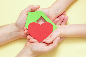 Obraz premium Close Up of dad and child hands holding green paper house with red heart on yellow background.