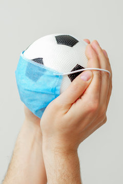 Closeup vertical image of soccer ball with a medical mask in male hands on a gray background. Cancellation of sporting events.