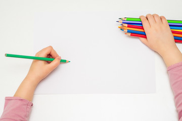 Top view of little hand of child drawing on blank paper by green pencil with bunch of colorful...