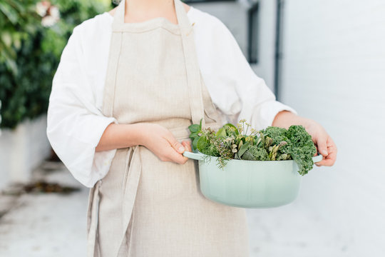 a woman with a linen apron and casserole filled with green vegetables and herbs