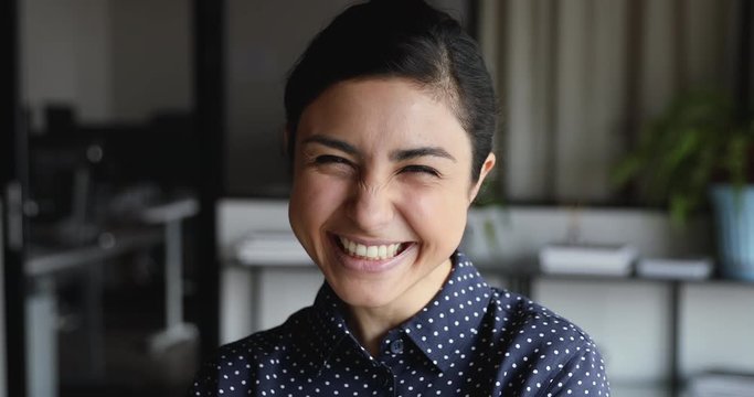 Cheerful funny young adult indian woman laughing looking at camera at home office. Happy pretty ethnic lady with healthy teeth, dental smile, having fun bursting into laughter indoor. Closeup portrait