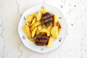 Grilled burgers and oven-roasted potatoes dish in a white plate isolated on white marble background. Top view, selective focus