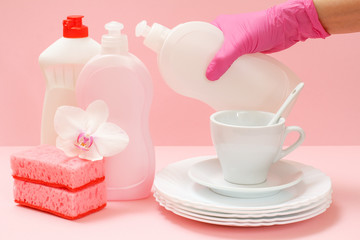 Woman's hand in protective glove with bottle of dishwashing liquid and utensil on a pink background.