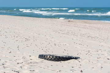 Big black rubber tire left on a sandy beach with blue sea and waves on background, environment pollution concept