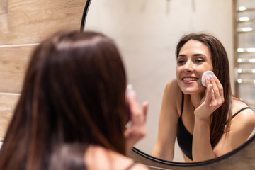 Beautiful young woman cleaning her skin with a cotton pad, looking at the mirror at home bathroom. Beauty, skin care concept, lifestyle