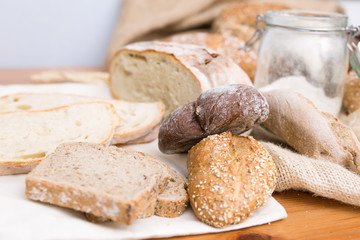 Buns, toasts and cut loafs on wooden table. Traditional wheat and rye bread, burlap sack and flour In glass jar. Closeup shot. Bakery or pastry concept