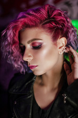 a girl with bright colored purple pink hair in a leather jacket black t shirt jeans in a dark dark bar against the background of neon signs poses leaned against the brick wall